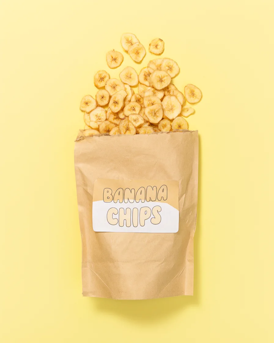 Top-down shot of a kraft paper bag labeled BANANA CHIPS with dried banana slices on a pastel yellow background.